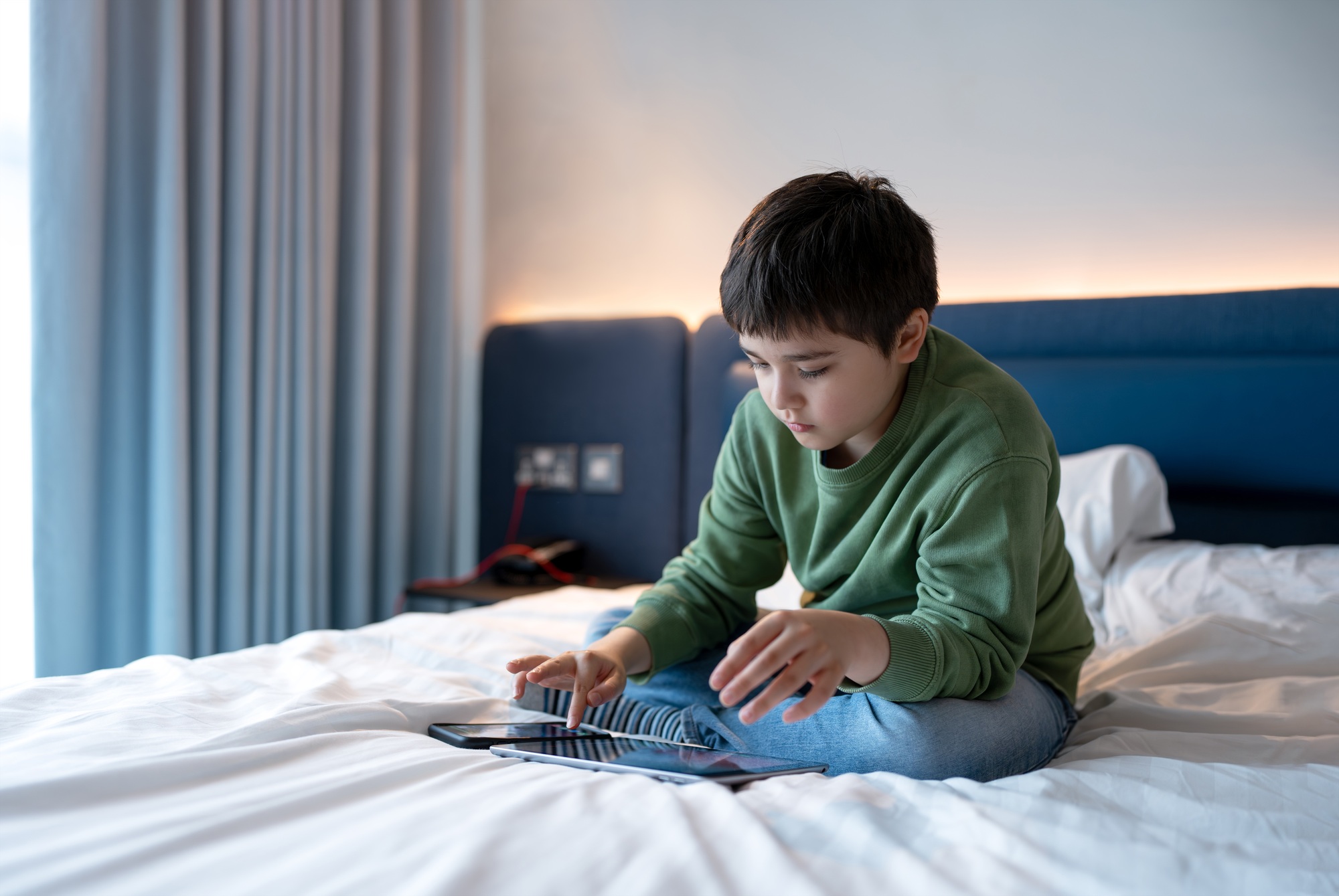 Kid boy using smartphone texting to friend,Child playing Game on Tablet sitting on a bed.